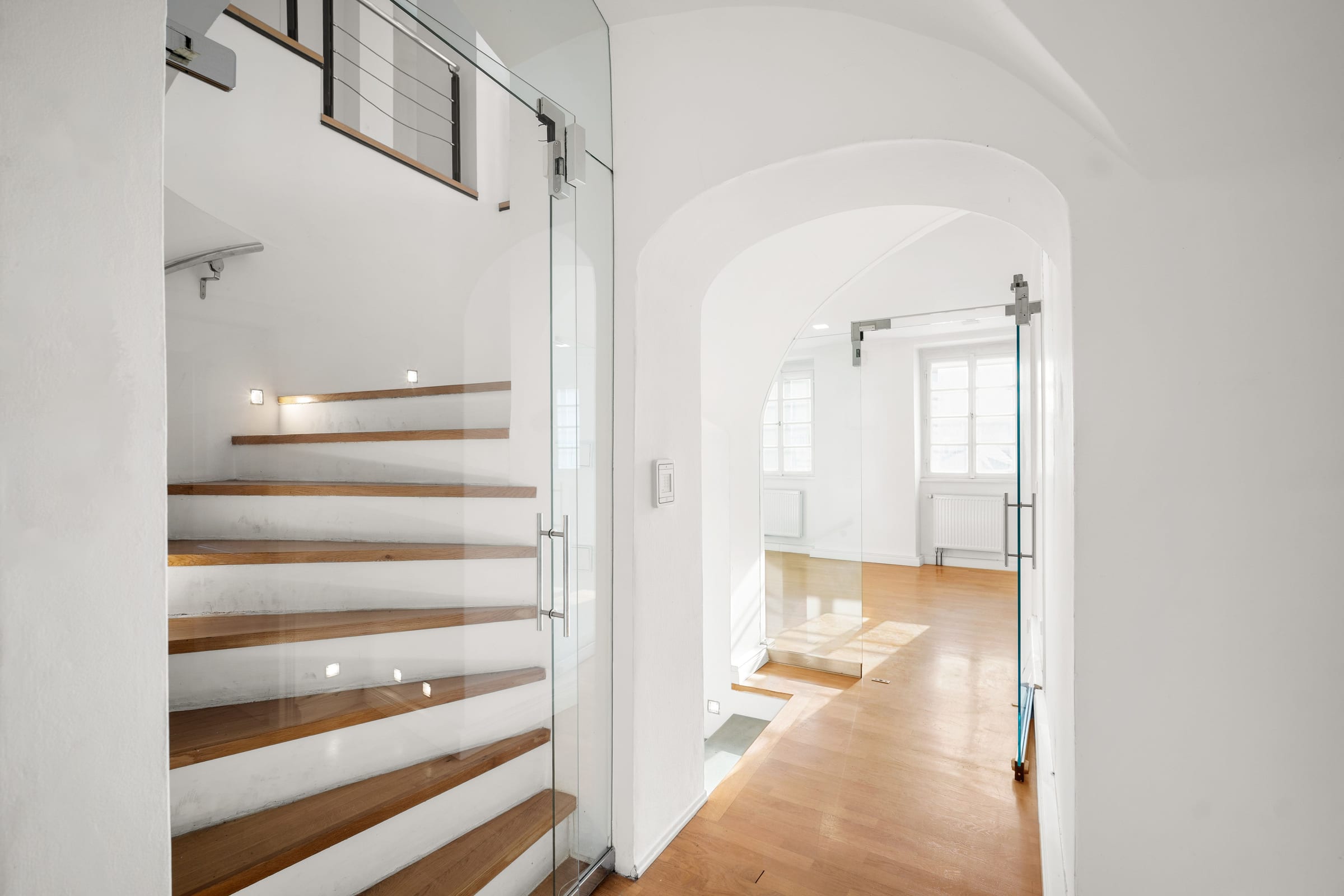 Treppe mit Holzstufen hinter einer Ganzglastür, dahinter ein gewölbter Durchgang